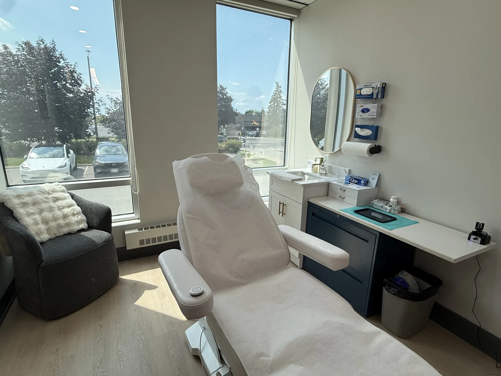 A treatment room at Artistry Clinic, with a reclining chair beside a window, an armchair for a companion, and a clean instrument counter.